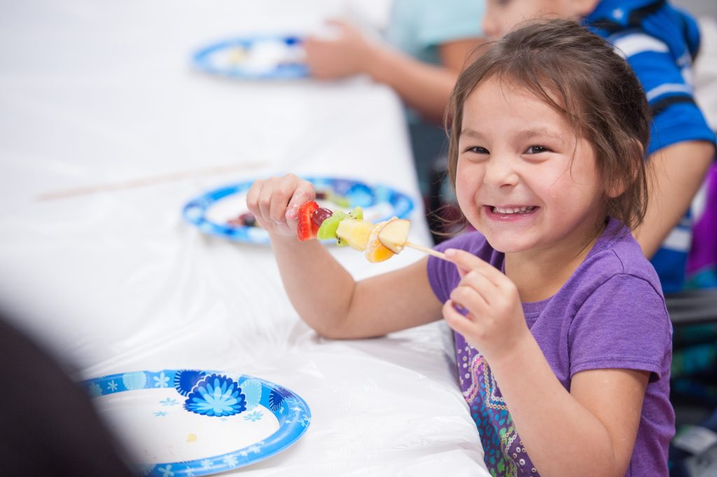 Little girl is smiling holding fruit on a stick. Children learn how to make fruit kebabs at school event.